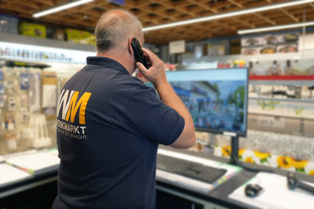 Ein Mann in blauem Poloshirt ist von hinten zu sehen, steht an einem Computer und telefoniert